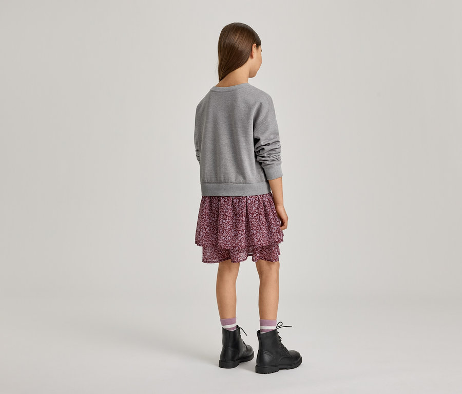 Mädchen von hinten, das einen grauen Pullover, einen gestuften Rock mit Blumenmuster, gestreifte Socken und schwarze Schnürstiefel trägt.