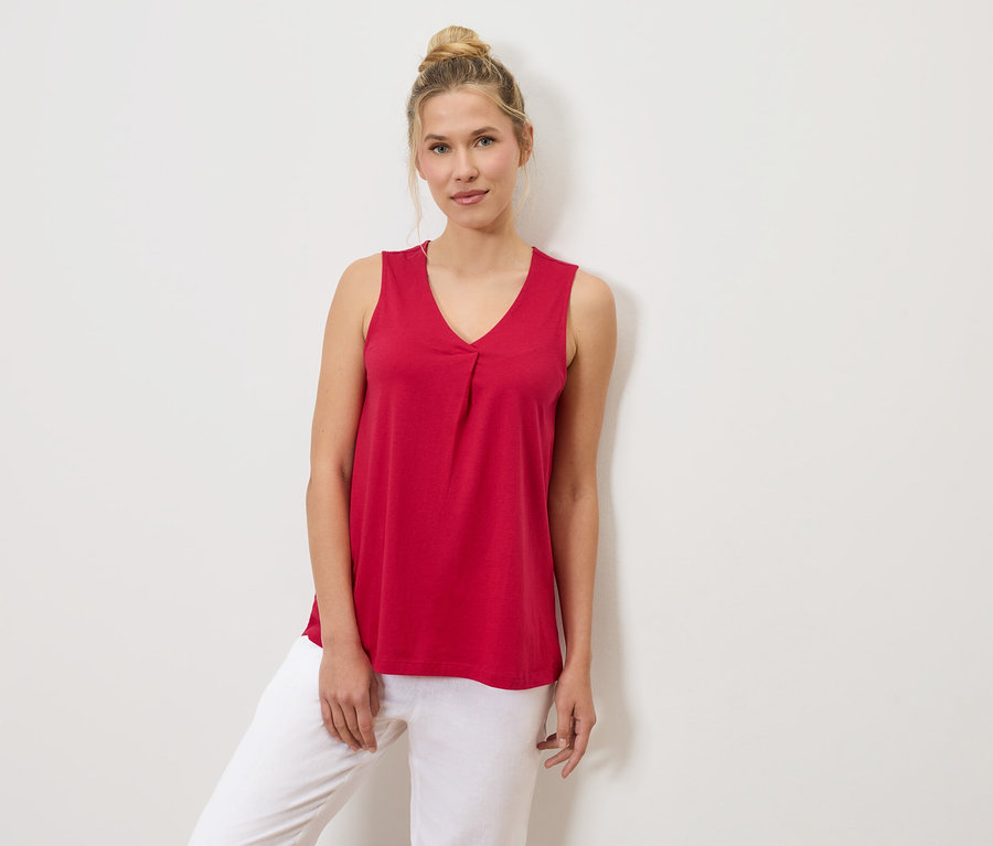 Frau mit Dutt trägt ein rotes Tanktop und weiße Hosen, die an einer weißen Wand lehnt.