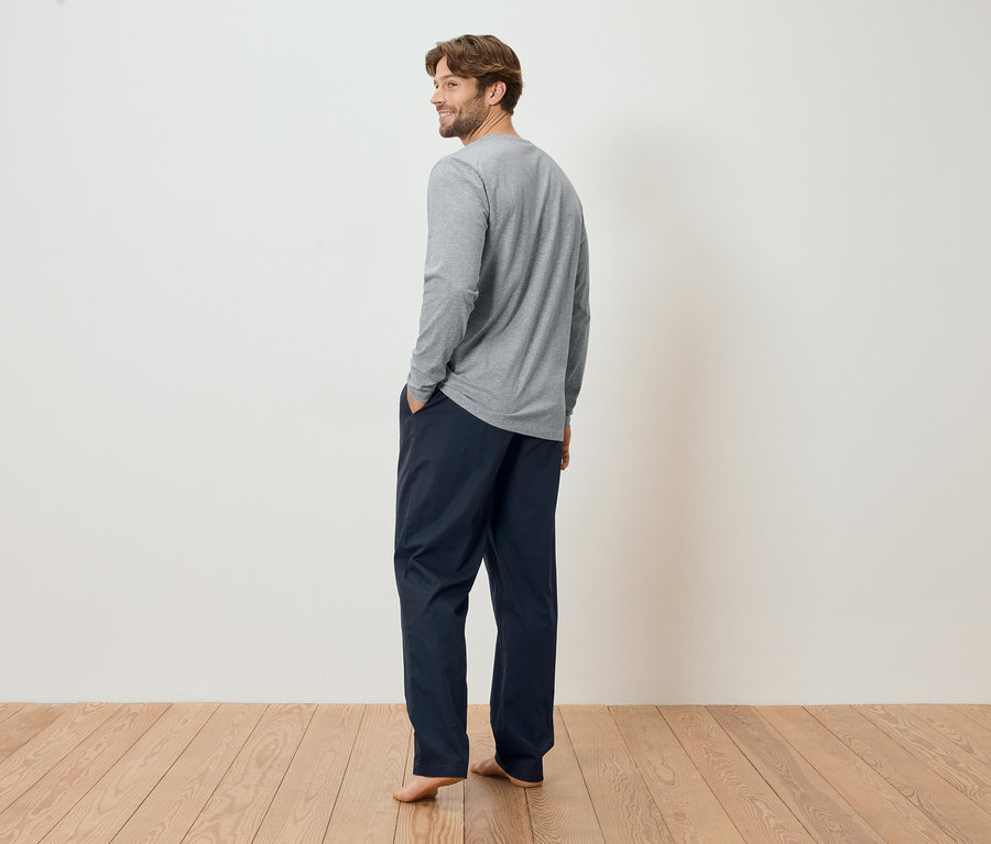 Ein Mann mit Bart trägt ein graues Langarmshirt und dunkelblaue Hosen, die Hände in den Taschen, mit Blick über die Schulter.