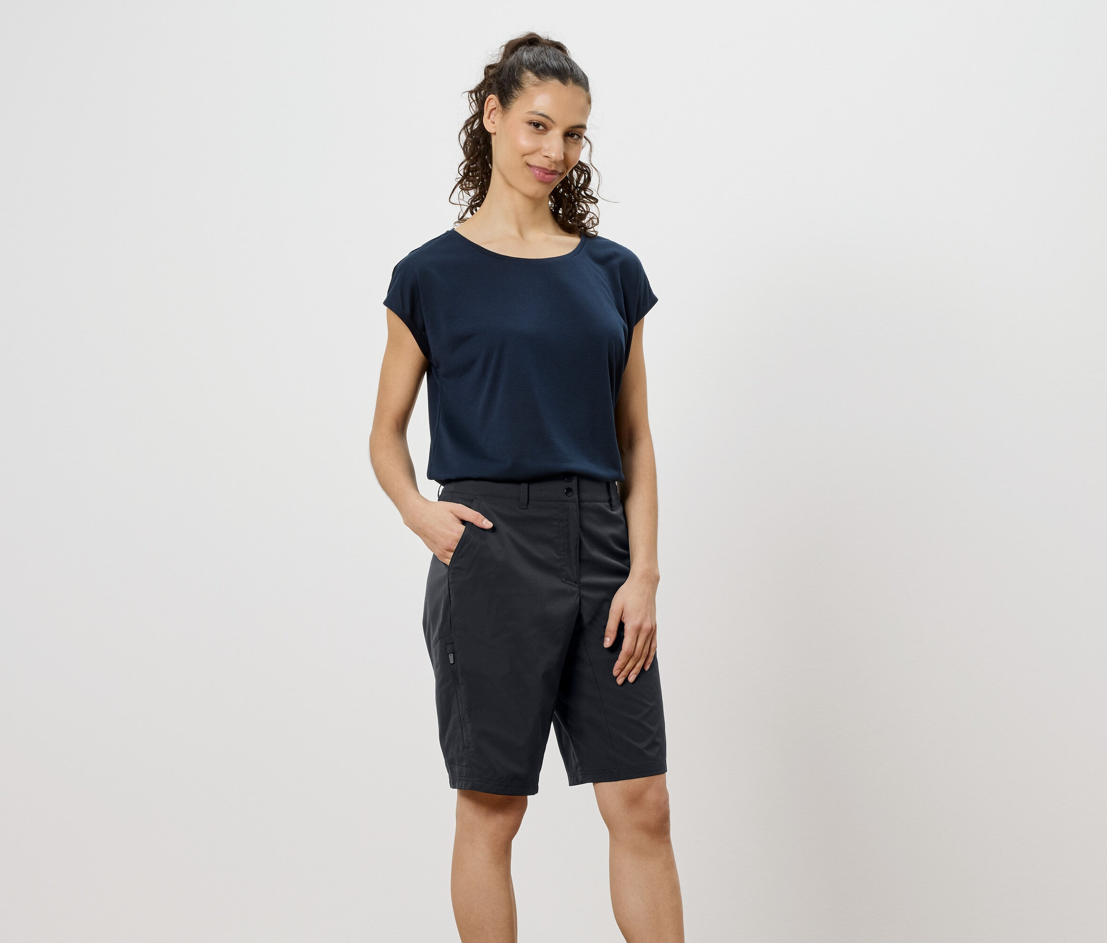 Frau mit lockigen, braunen Haaren, die ein marineblaues T-Shirt und graue Shorts trägt und die Hand in der Hosentasche hat.