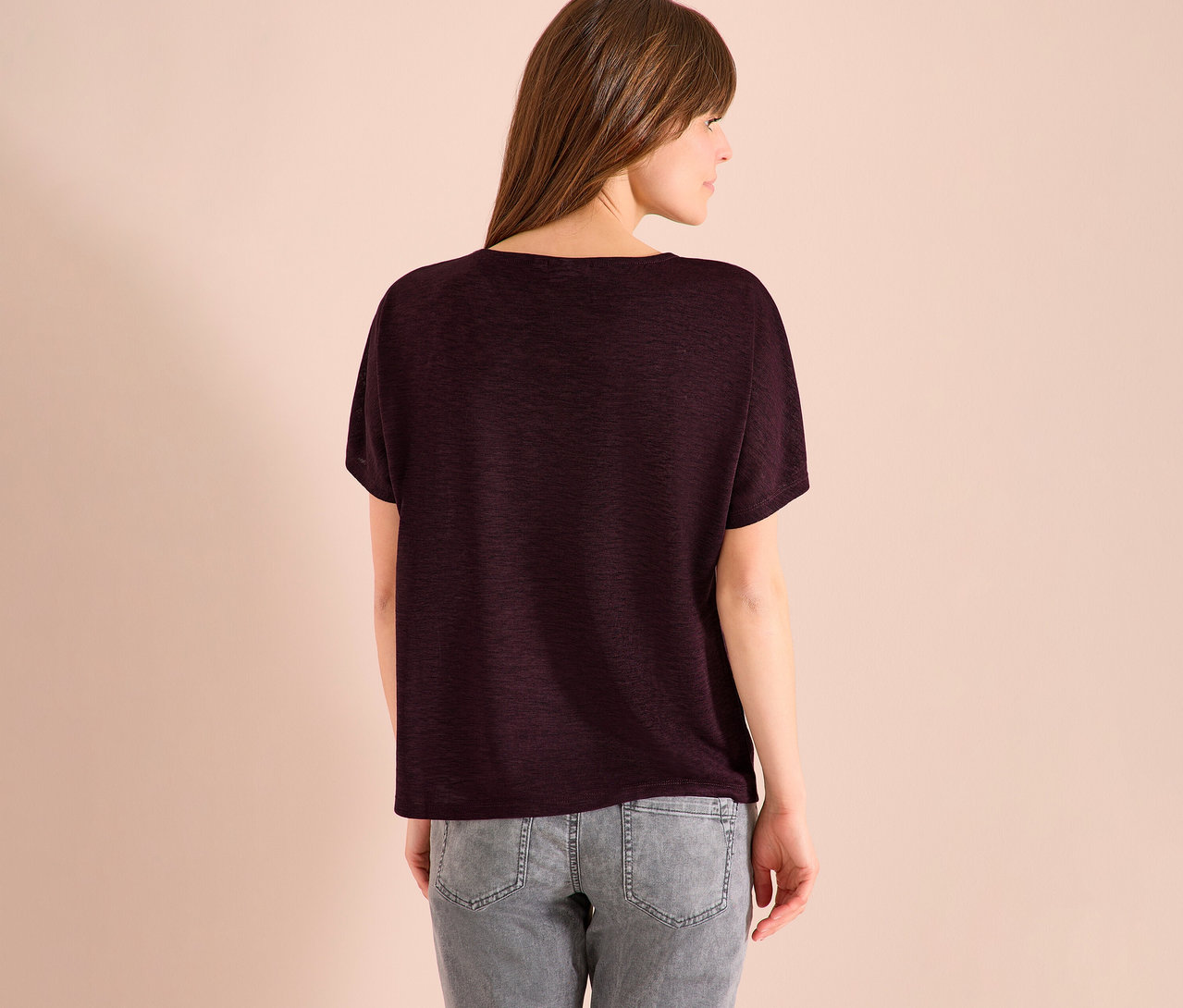 Rückansicht einer Frau mit braunen Haaren, die ein dunkelrotes T-Shirt und graue Jeans trägt.