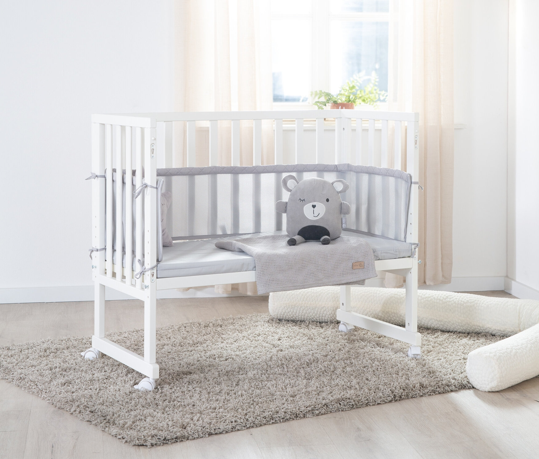 Weißes roba 3-in-1-Stuben-Beistellbett mit grauer Polsterung und Bär-Stofftier auf einem grauen Teppich.