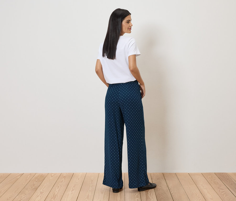 Frau mit weißem T-Shirt und blau-weiß gepunkteter Hose, die über einem Holzboden steht und über die Schulter blickt.