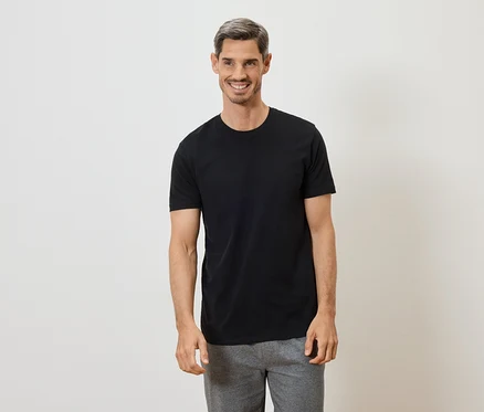 Mann mit grau melierter Frisur lächelt in schwarzem T-Shirt und grauer Hose vor weißem Hintergrund.