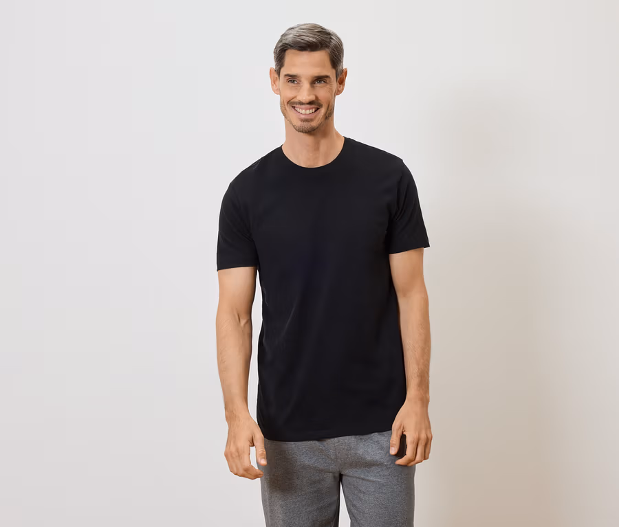 Mann mit grau melierter Frisur lächelt in schwarzem T-Shirt und grauer Hose vor weißem Hintergrund.