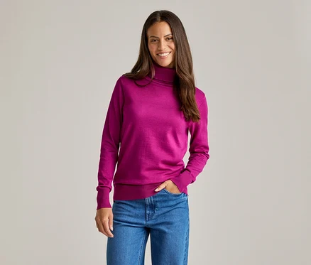Frau lächelt und trägt einen pinken Rollkragenpullover und blaue Jeans, eine Hand in der Hosentasche.