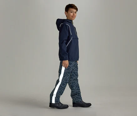 Ein Junge in marineblauer Regenjacke mit Kapuze und gemusterter Regenhose mit reflektierendem Streifen geht seitlich nach rechts.