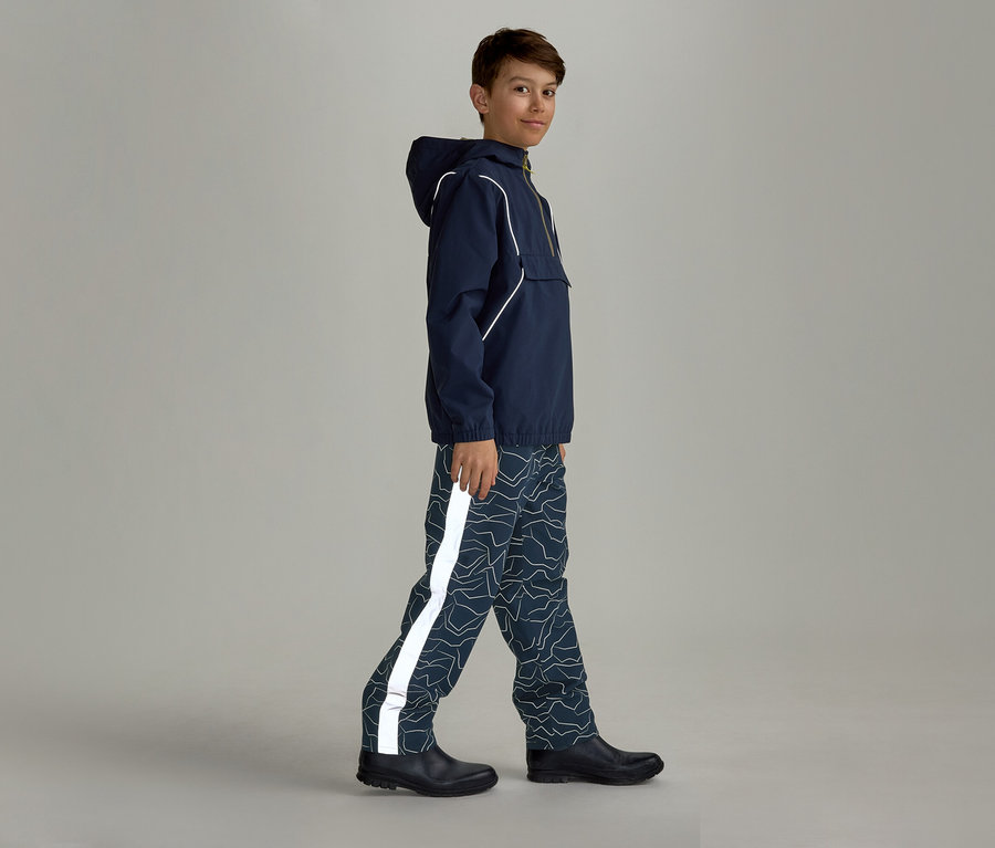 Ein Junge in marineblauer Regenjacke mit Kapuze und gemusterter Regenhose mit reflektierendem Streifen geht seitlich nach rechts.