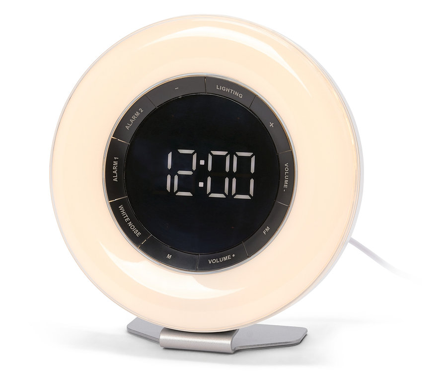 Ein weißer, runder Lichtwecker mit einem beleuchteten äußeren Ring und einem dunklen zentralen Display, das "12:00" anzeigt. Steuerungstasten wie "ALARM 1", "ALARM 2", "LIGHTING", "WHITE NOISE", "FM" und "VOLUME" sind um das Display herum sichtbar. Der Wecker steht auf einem silbernen Sockel.