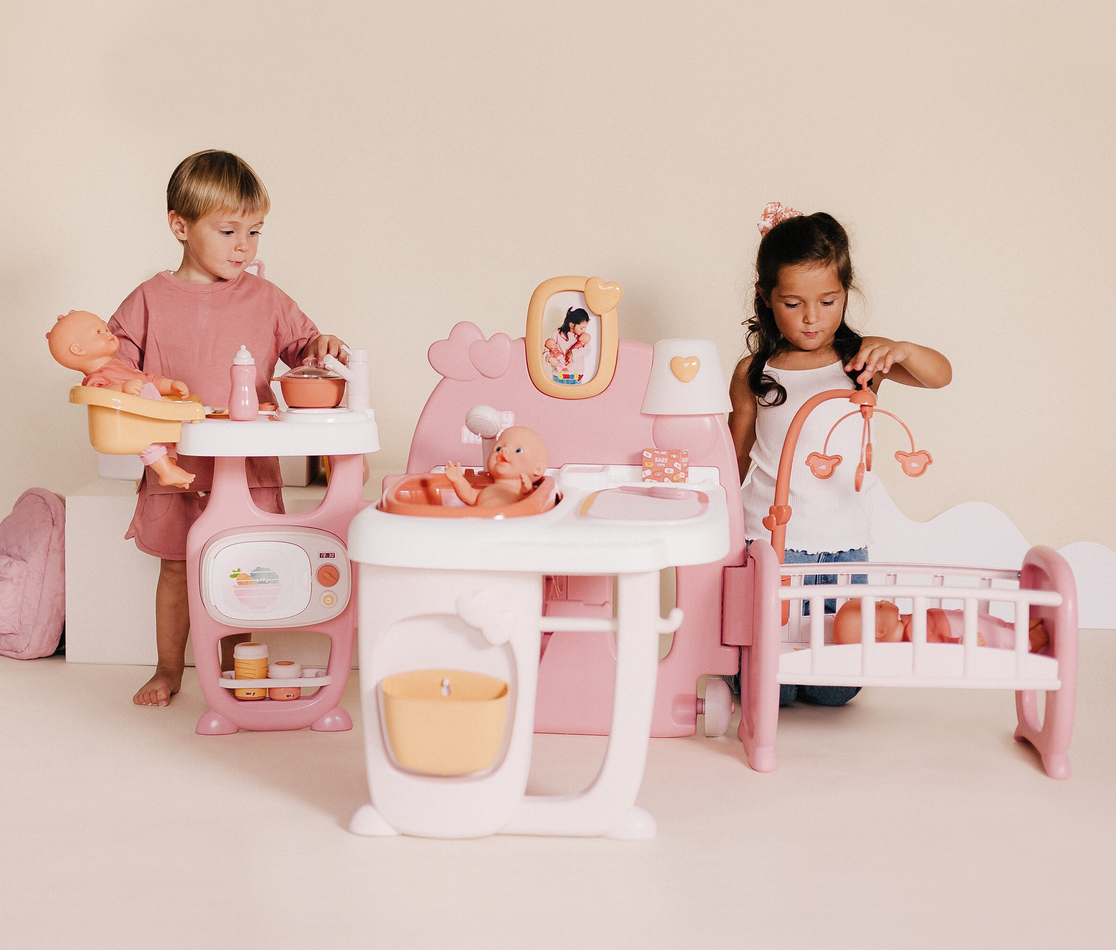 Zwei Kinder spielen mit einem Smoby Baby Nurse Puppen-Spielcenter, das eine Puppenküche, ein Bett und eine Badestation umfasst.