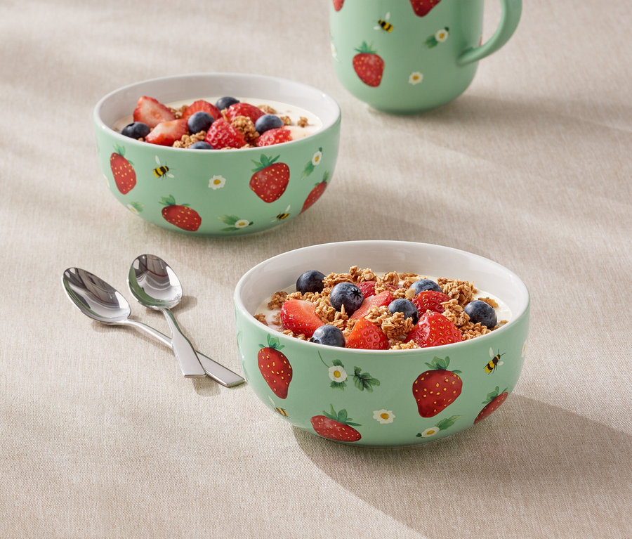 Zwei mit Joghurt, Erdbeeren und Blaubeeren gefüllte Schalen mit Erdbeermuster und ein Krug stehen auf einem Tisch.