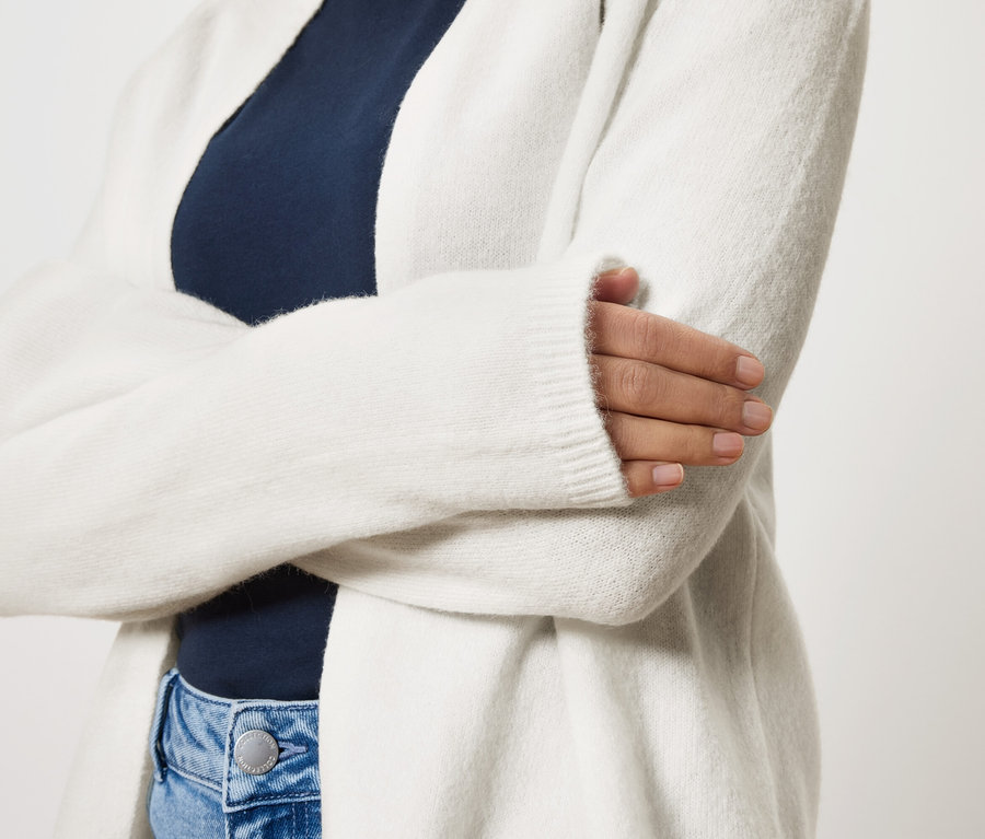 Frau mit verschränkten Armen trägt eine lange, weiße Strickjacke über einem marineblauen Oberteil und blauen Jeans.