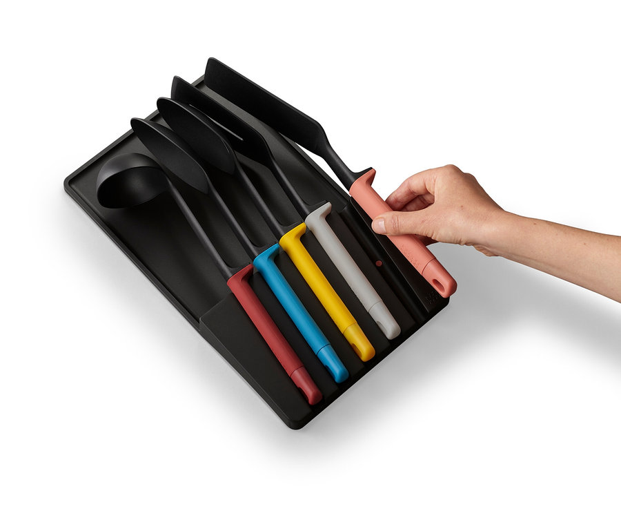 Eine Hand legt ein Küchenutensil mit rotem Griff in einen schwarzen Schubladeneinsatz. Weitere Küchenutensilien sind ebenfalls im Einsatz.