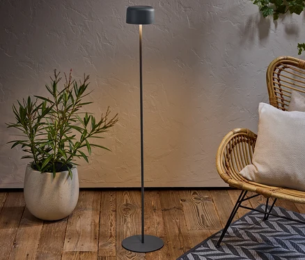 Eine Stehlampe steht neben einer Topfpflanze und einem Korbstuhl mit Kissen auf einem gemusterten Teppich.
