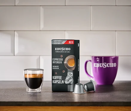 Eine schwarze Packung EDUSCHO Espresso Intensiv neben einem Espressoglas, einer lila Tasse und zwei Kapseln.