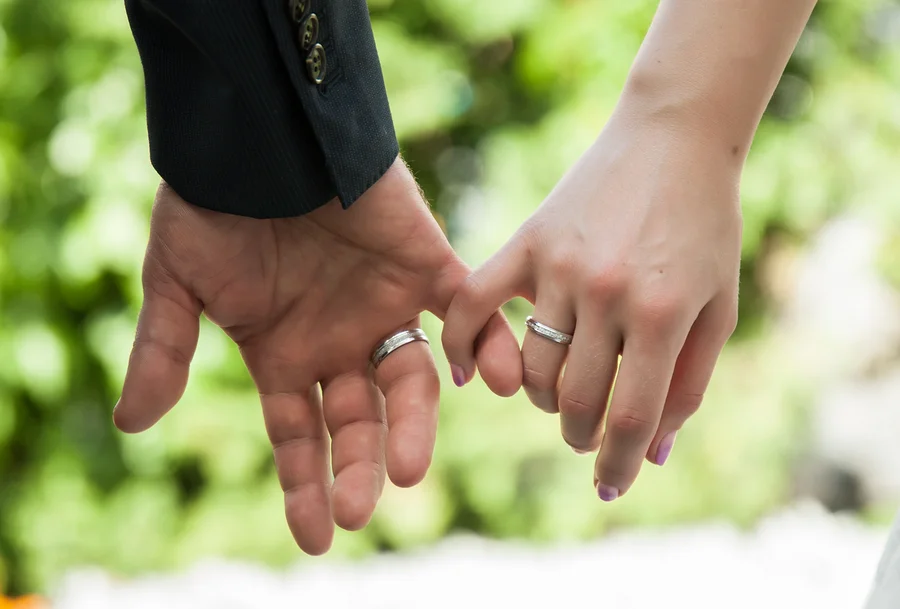 Eine Männerhand und eine Frauenhand halten sich an den kleinen Fingern, beide tragen silberne Ringe. Die Männerhand links trägt einen dunklen Anzugärmel, die Frauenhand rechts hat hellvioletten Nagellack. Im Hintergrund ist verschwommenes grünes Laub zu sehen.