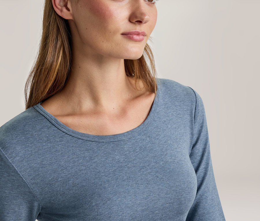 Eine Frau mit langen braunen Haaren trägt ein blaues Langarmshirt mit Rundhalsausschnitt.