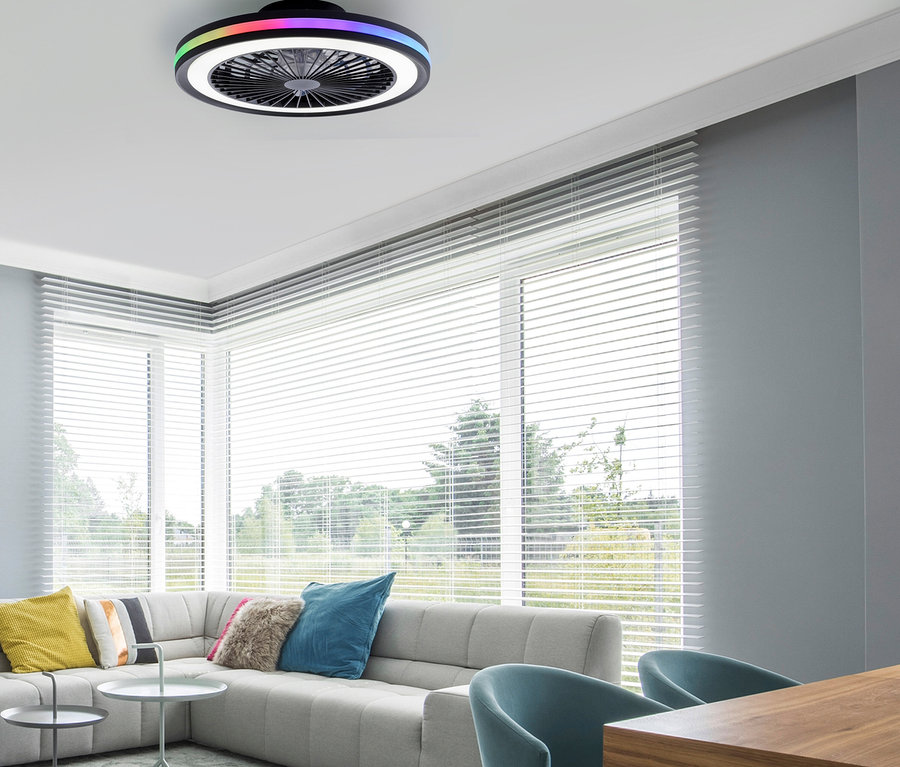 Ein modernes Wohnzimmer mit einer schwarzen Deckenventilatorleuchte mit bunten und weißen LED-Ringen. Ein hellgraues Ecksofa mit bunten Kissen, zwei hellblauen Sesseln und einem Holztisch sind sichtbar. Große Fenster mit weißen Jalousien blicken auf eine grüne Landschaft.