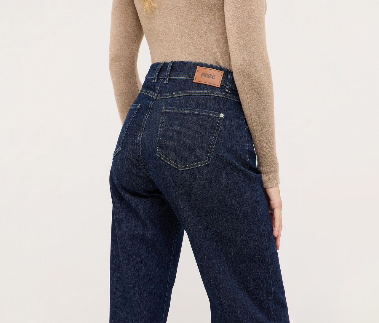 Rückenansicht einer Person in dunkelblauen Jeans und einem beigen Langarmshirt. Die Jeans hat einen braunen 'ANGELS'-Markenpatch am hinteren Bund.