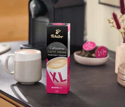 Eine Kaffeemaschine steht neben einer Packung Caffè Crema Wake Up XL – 80 Kapseln und einer Tasse auf einer Küchentheke.