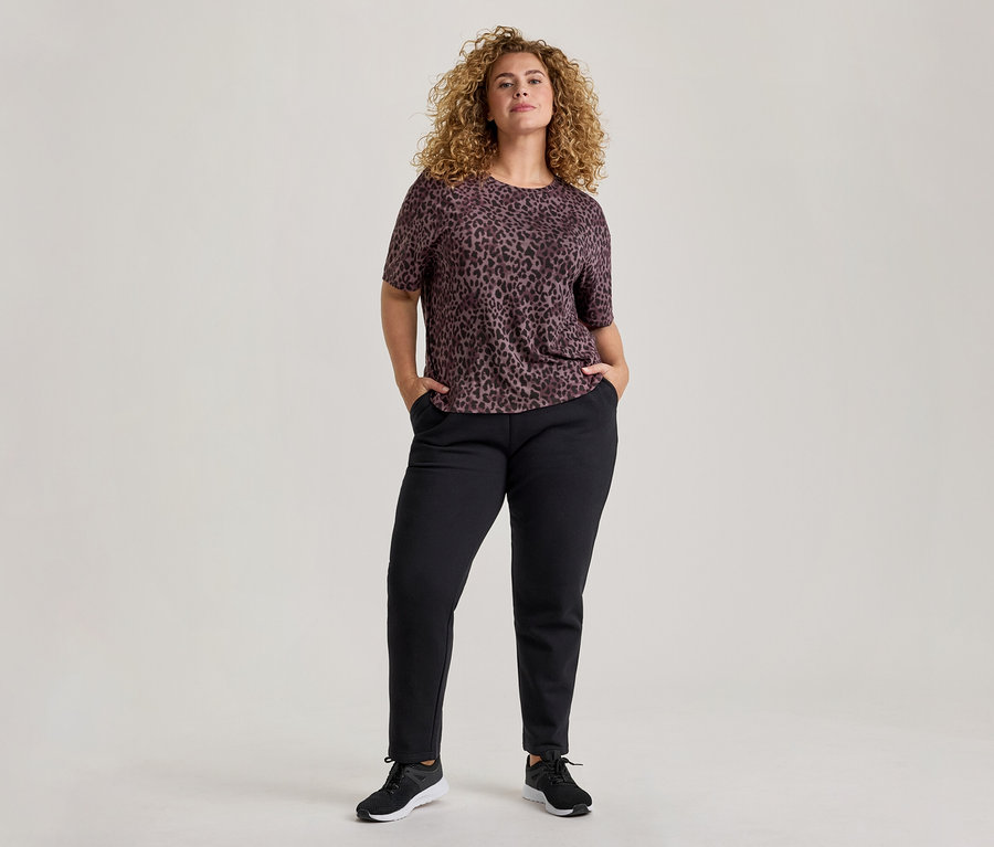 Eine Frau mit lockigem Haar steht frontal, trägt ein kurzärmeliges, dunkelviolettes Leopardenmuster-Oberteil, schwarze Hosen und schwarze Turnschuhe. Ihre Hände stecken in den Hosentaschen.