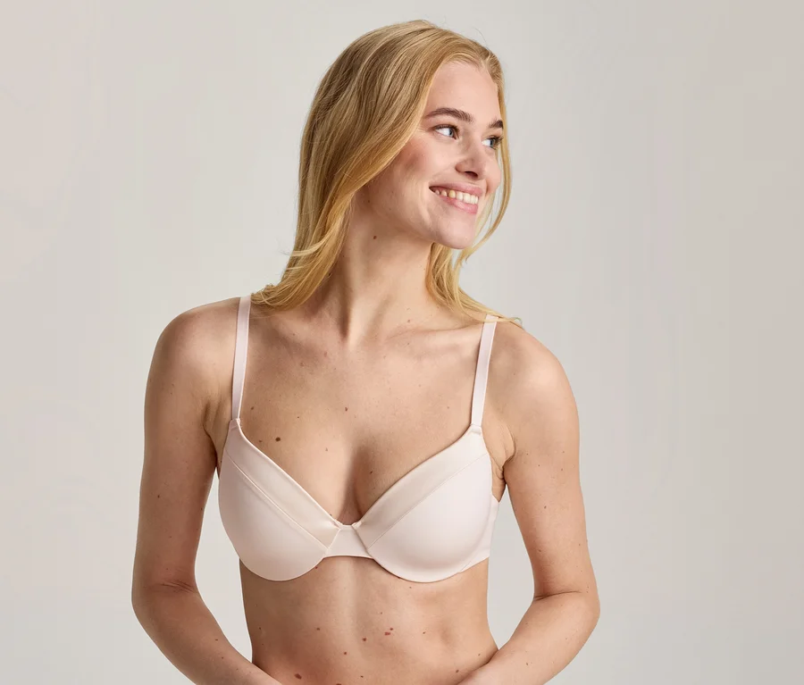 Blonde Frau trägt einen hellrosa BH, lächelt und blickt nach rechts.