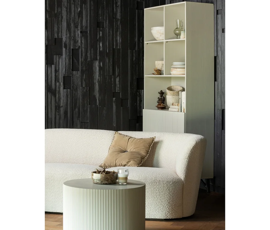 Ein modernes Wohnzimmer mit einem cremefarbenen Bouclé-Sofa mit braunem Kissen, einem runden geriffelten Couchtisch mit Schale und Glas sowie einem hellen Regal vor einer dunklen Holzwand.