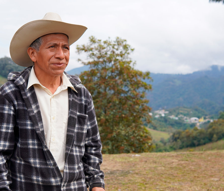 Ein älterer Mann mit hellem Cowboyhut und schwarz-weiß karierter Jacke steht im Freien und blickt nach rechts. Im Hintergrund sind grüne Berge, ein Tal mit einem Dorf und ein bewölkter Himmel zu sehen.