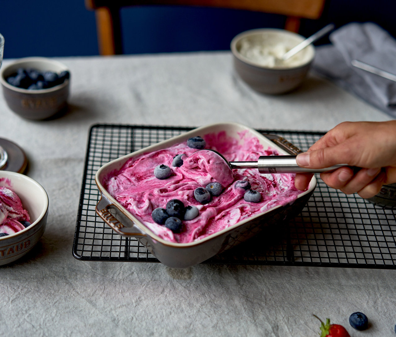 Eine Hand schöpft pink-weißes Beeren-Eis aus einer antikgrauen STAUB Auflaufform, die mit Heidelbeeren belegt ist und auf einem Gitterrost steht. Weitere Schüsseln mit Dessert und Heidelbeeren stehen auf einem hellen Tischtuch.