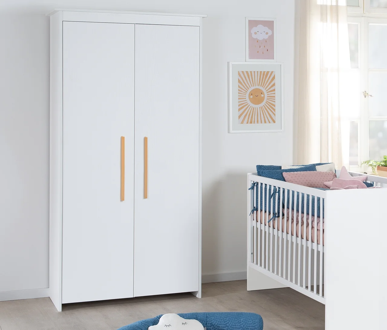 Ein weißer zweitüriger Kleiderschrank mit Holzgriffen steht neben einem weißen Babybett mit blauer und rosa Bettwäsche. Zwei gerahmte Bilder, eines mit Sonne und eines mit Wolke, hängen an der Wand über dem Bett.