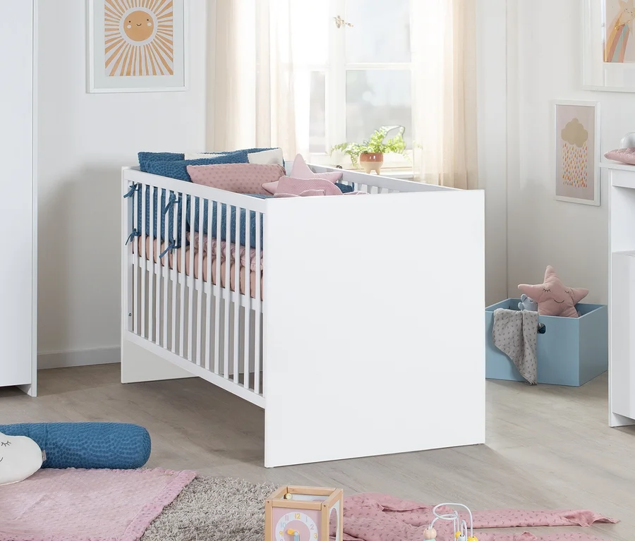 Ein helles Kinderzimmer mit einem weißen Babybett, gefüllt mit blauer und rosa Bettwäsche und einem Sternkissen. Dahinter ist ein Fenster mit Vorhängen. Sonnen-, Regenbogen- und Wolkenbilder schmücken die Wände. Am Boden liegen ein Teppich, eine blaue Aufbewahrungsbox und Babyspielzeug.