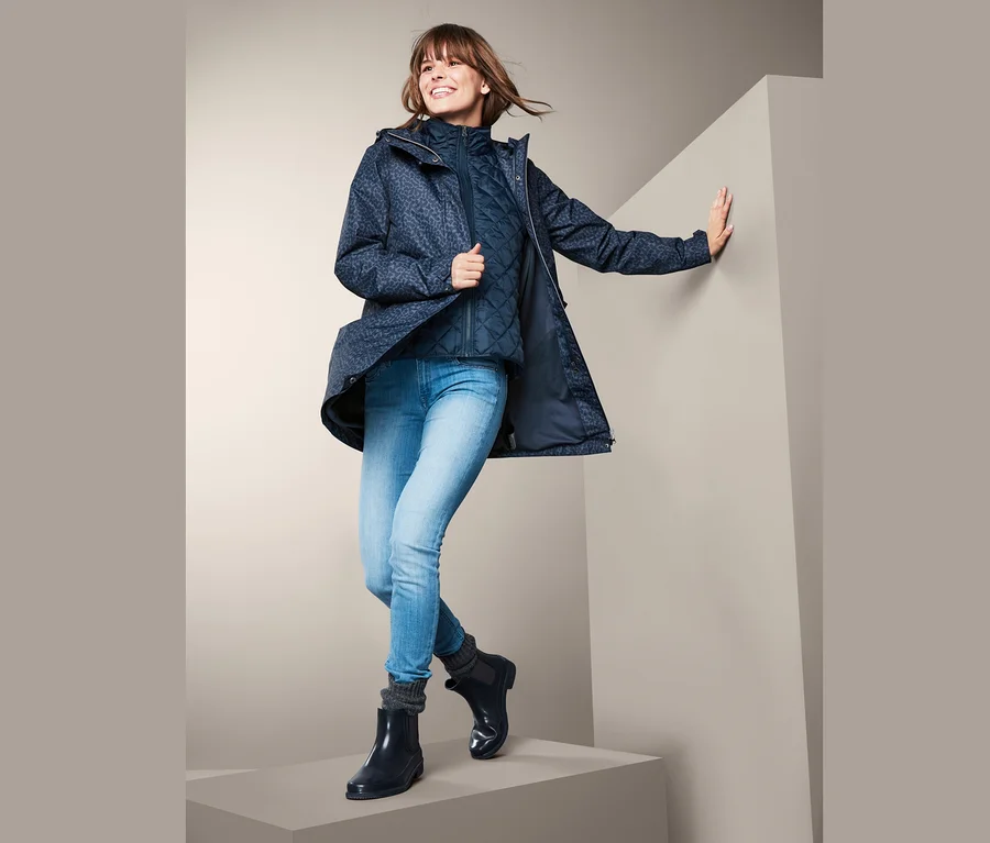 Eine lächelnde Frau posiert in einer 3-in-1-Regenjacke, einer blauen Steppweste, Jeans und Gummistiefeln.