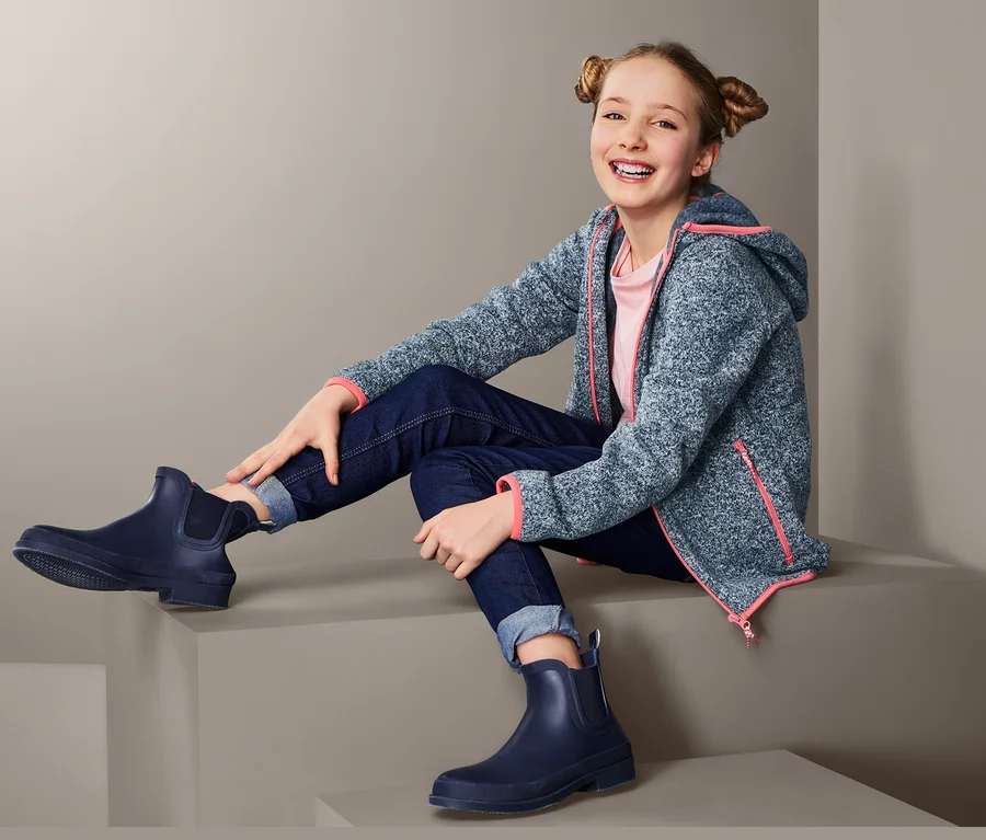 Ein Mädchen sitzt auf einer Stufe und trägt eine Strickfleecejacke mit Kapuze, Jeans und Gummistiefel.