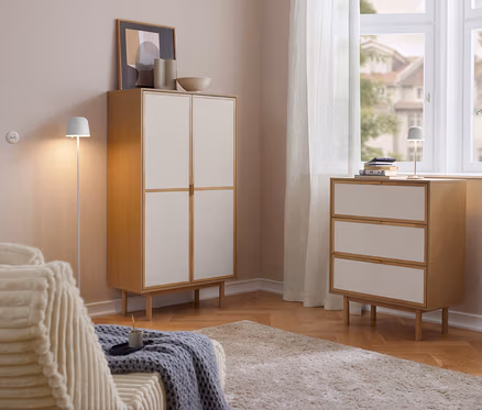 Helles Wohnzimmer mit einem Cabinetschrank mit 2 Türen und einer Kommode mit 3 Schubladen aus Holz und hellen Fronten. Auf der Kommode steht eine Eglo LED-Akku-Tischleuchte »Mannera«. Links ein Sofa, rechts ein Fenster mit Vorhängen.