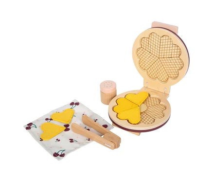Ein offenes small foot Waffeleisen-Set "tasty" in Rot mit zwei gelben Herzwaffeln. Daneben liegen eine weitere Herzwaffel, eine Holzzange und ein Streuer auf einem Kirschmuster-Tuch.