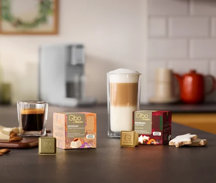 Eine Qbo ICONIC Light Gold Kaffeemaschine im Hintergrund. Auf einer Küchenzeile stehen zwei Qbo Espresso Kapselpackungen, White Choc & Mandel und Zimtstern, mit passenden Kapseln, Espresso und Latte Macchiato, Mandeln, weißer Schokolade und Zimtsternen.