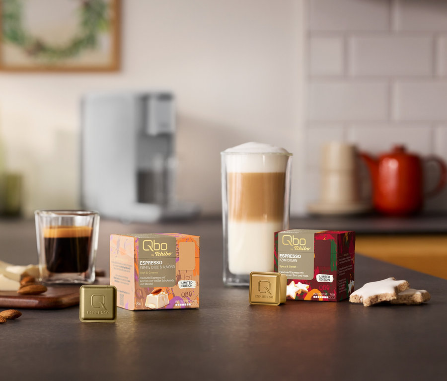 Eine Qbo ICONIC Light Gold Kaffeemaschine im Hintergrund. Auf einer Küchenzeile stehen zwei Qbo Espresso Kapselpackungen, White Choc & Mandel und Zimtstern, mit passenden Kapseln, Espresso und Latte Macchiato, Mandeln, weißer Schokolade und Zimtsternen.