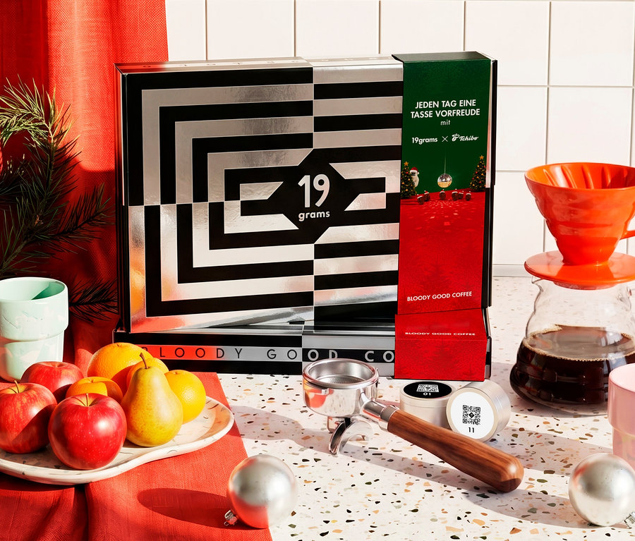 Ein schwarz-silberner Kaffee-Adventskalender mit geometrischem Muster und der Aufschrift „JEDEN TAG EINE TASSE VORFREUDE mit 19grams X Tchibo“ steht auf einer Theke. Daneben liegen Äpfel, Birnen und Orangen auf einem Teller, ein Pour-over-Kaffeebereiter mit Glaskanne, zwei Tassen, ein Siebträger, Kaffeekapseln und Weihnachtsbaumkugeln.