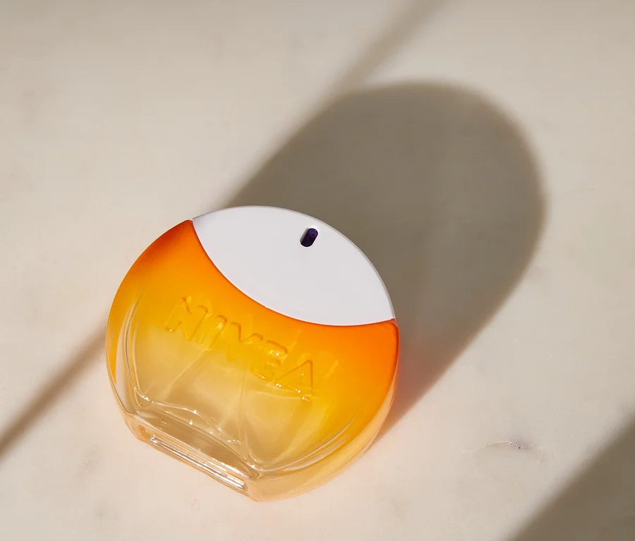 Eine ovale Nivea-Flasche mit orange-gelbem Farbverlauf und weißer Kappe liegt auf einer hellen Marmoroberfläche und wirft einen großen Schatten.