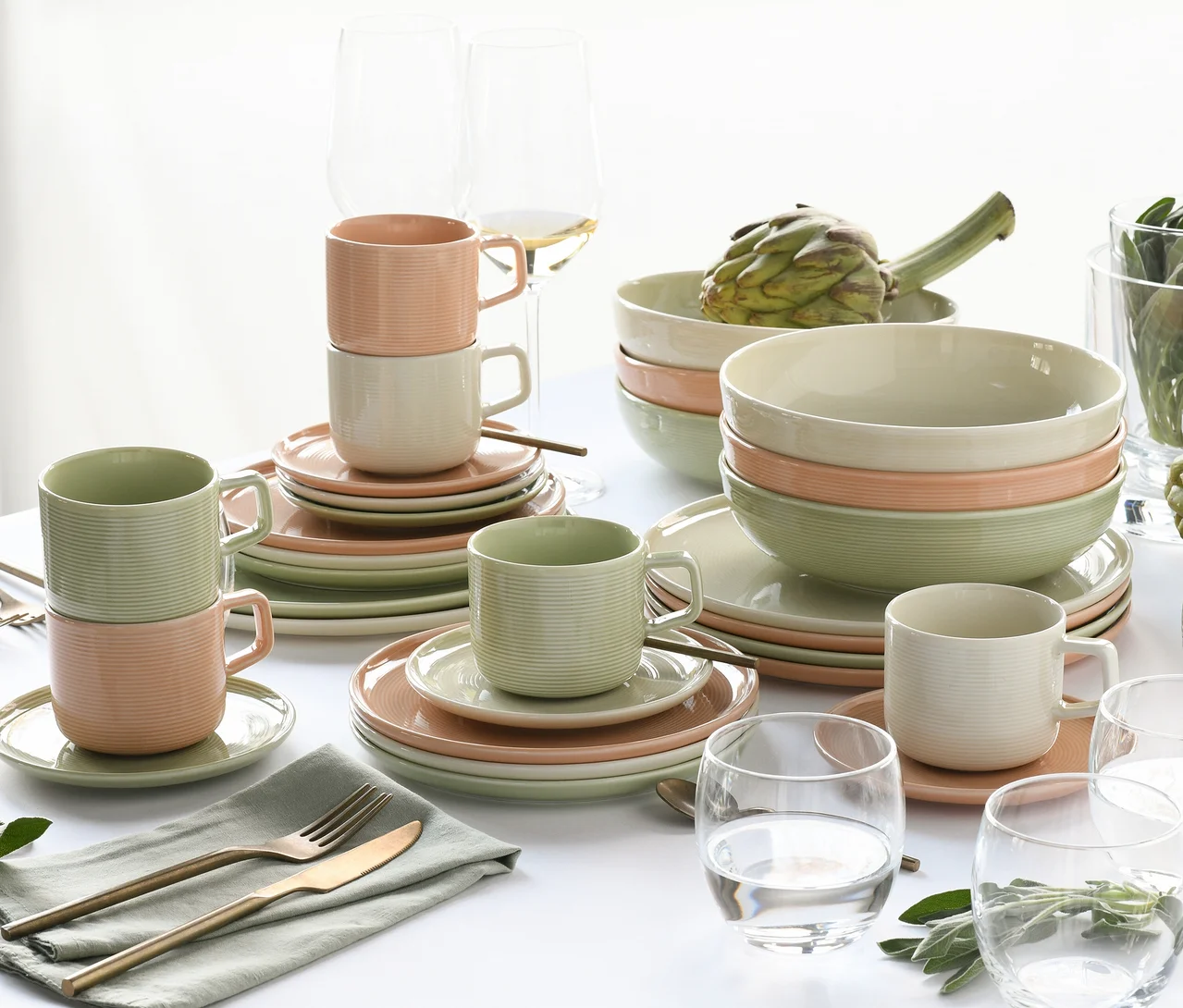 Ein Tisch, gedeckt mit einem 30-teiligen creaTable Service "Session Romance", bestehend aus gerippten Tellern, Schalen und Tassen in Hellgrün, Hellrosa und Creme. Weingläser, Wassergläser und Besteck auf einer grünen Serviette sind ebenfalls zu sehen, mit einer Artischocke in einer Schale.