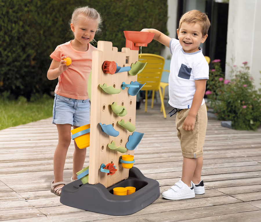 Ein Junge und ein Mädchen spielen auf einer Holzterrasse mit einer Smoby Life Activity Wall. Der Junge gießt Wasser oben hinein, während das Mädchen einen gelben Ball und einen Eimer hält.