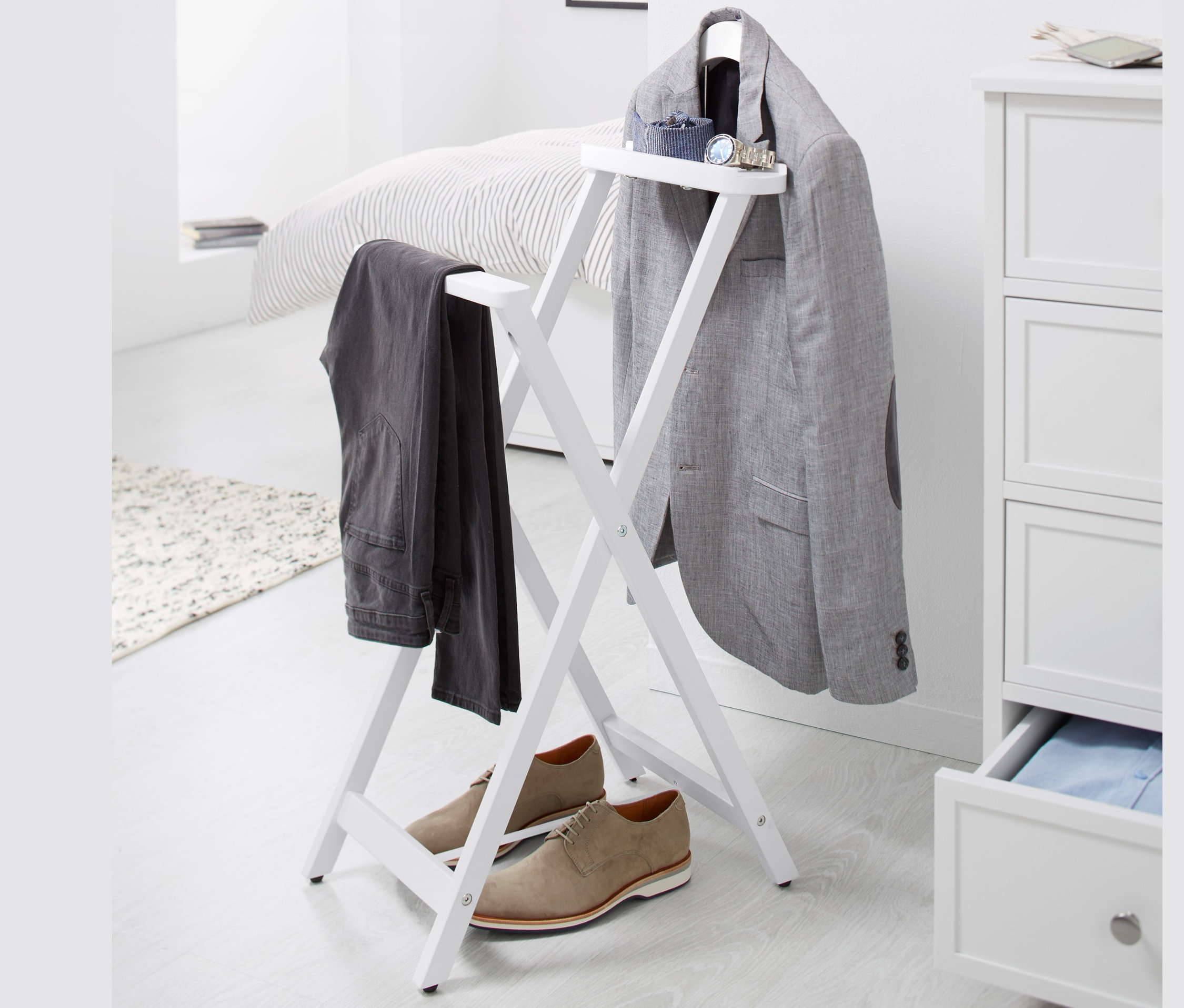 Auf einem weißen Hintergrund steht ein stummer Diener, an dem ein grauer Blazer und eine Hose hängen, darunter stehen braune Schuhe.