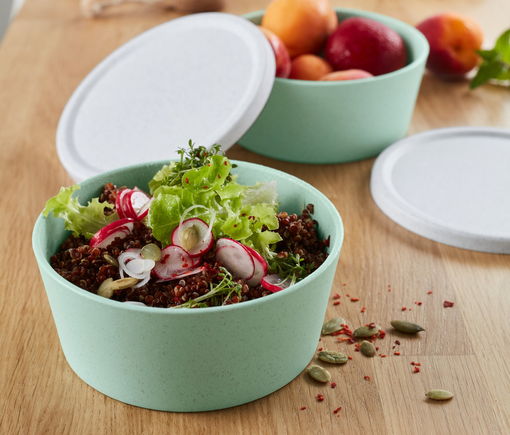 Zwei hellgrüne KOZIOL-Schalen mit weißen Deckeln. Eine Schale enthält Salat und Radieschen, die andere Aprikosen und Pflaumen.