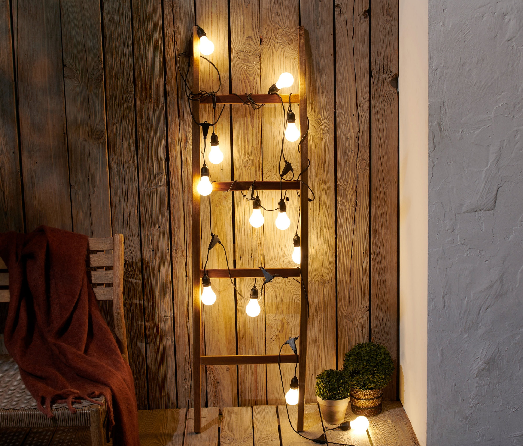 Eine LED-Outdoor-Lichterkette ist um eine Holzleiter gewickelt, die an einer Holzwand lehnt.