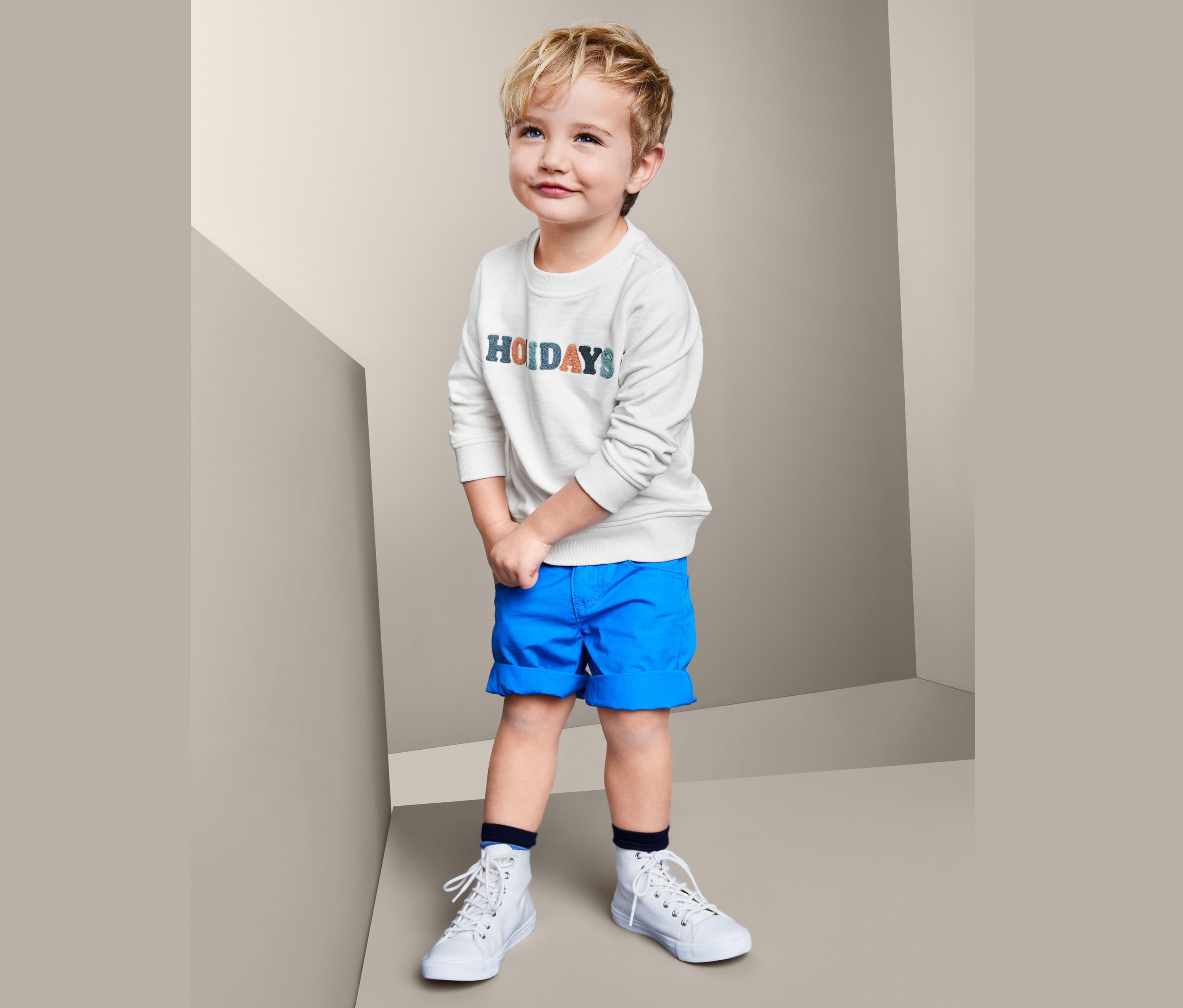 Junge trägt Baumwoll-Sweatshirt mit der Aufschrift Holidays, blaue Shorts und weiße Turnschuhe, steht in einer Ecke des Raumes.