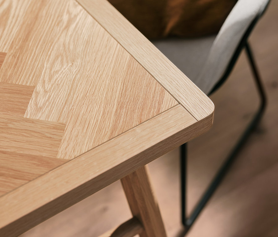 Detail einer Holztischplatte mit Fischgrätmuster und einem beige gepolsterten Schalenstuhl mit Metallbeinen.