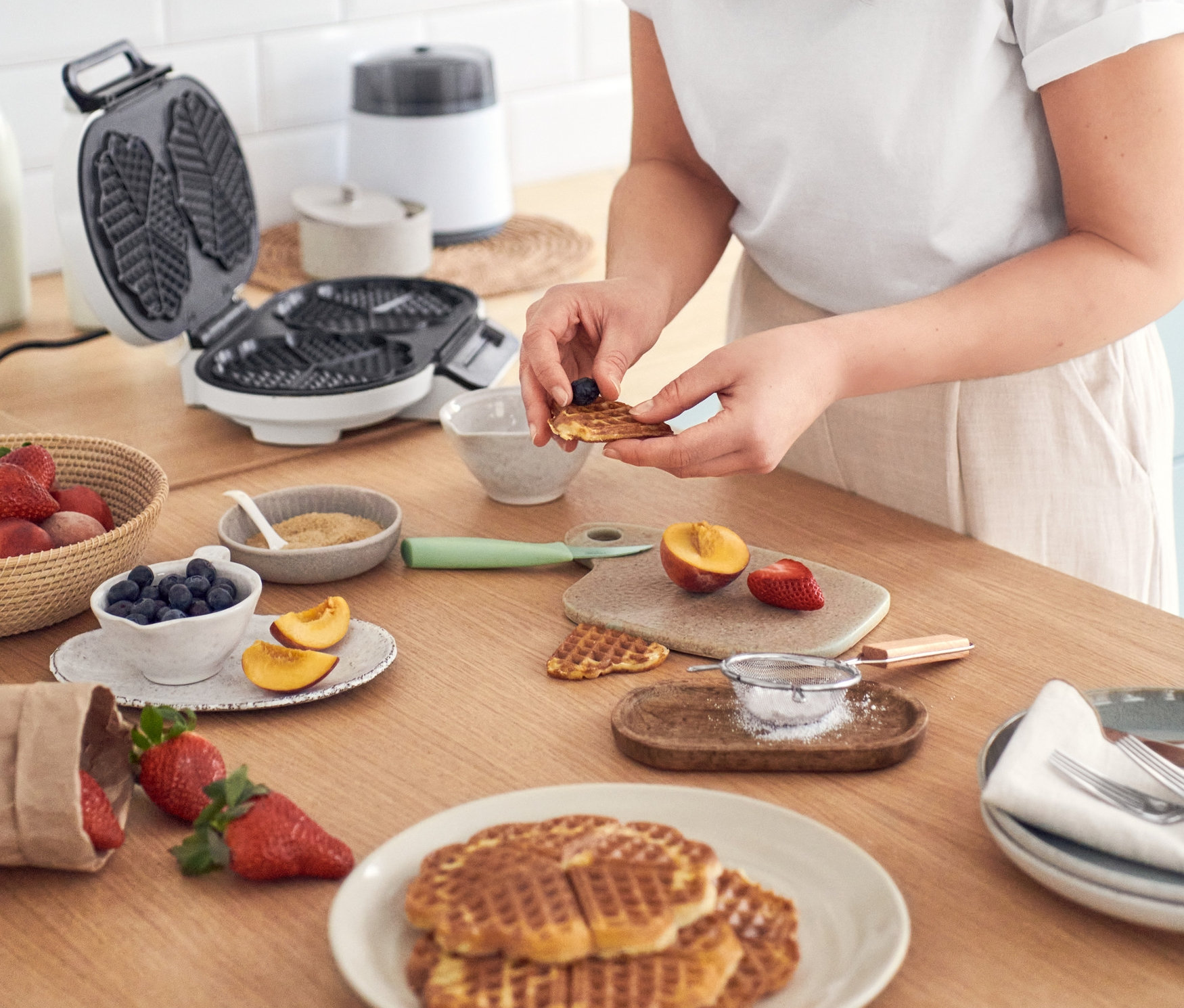 Frau dekoriert Waffeln mit Blaubeeren an einem Tisch mit Waffeln, einer Eismaschine »Comfortice« und dem WILFA Waffeleisen »TRADITION DOUBLE WDWA-516W«.
