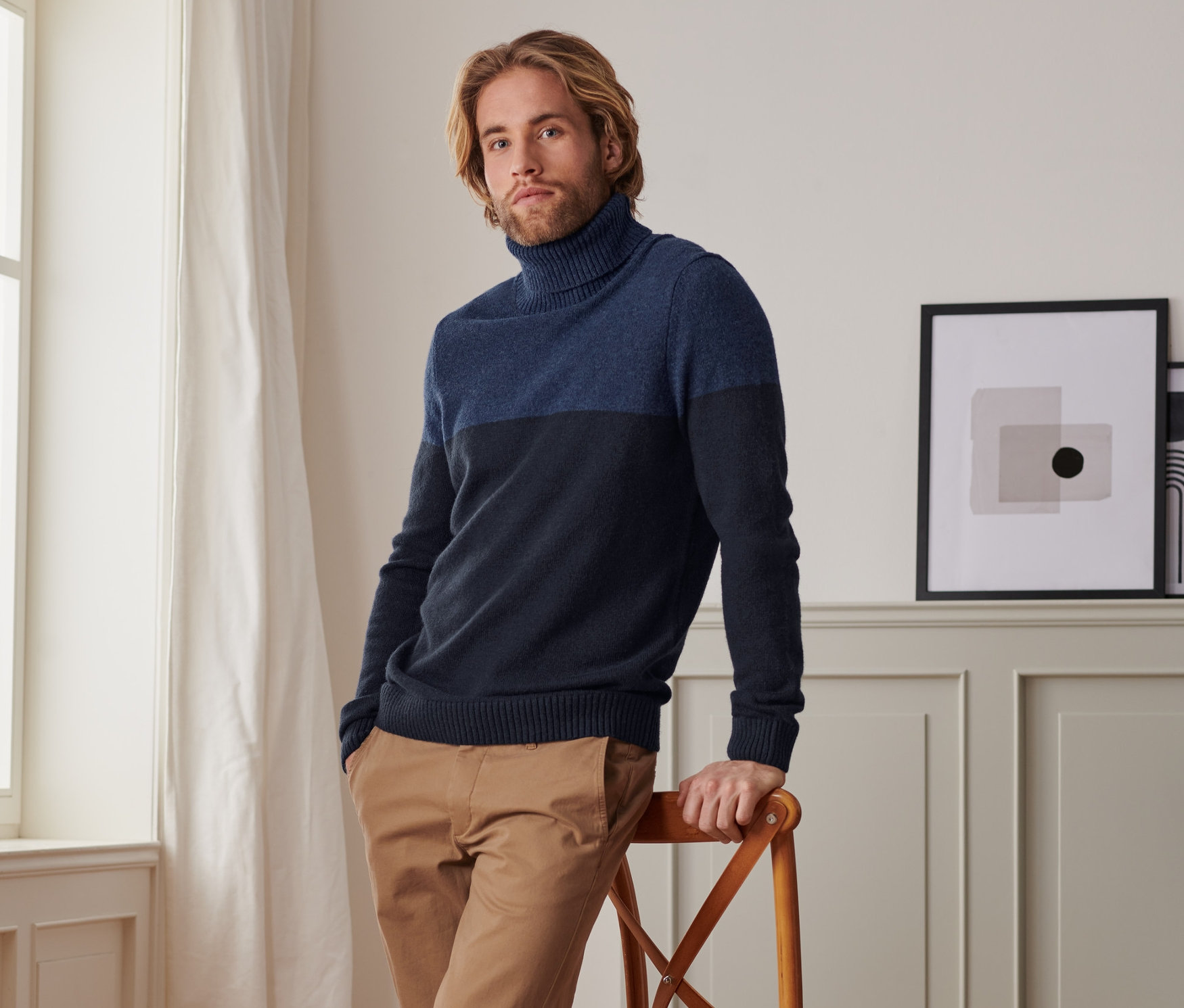 Mann, der sich an einen Stuhl lehnt, trägt einen Rollkragen-Pullover, braune Hose und weiße Turnschuhe.