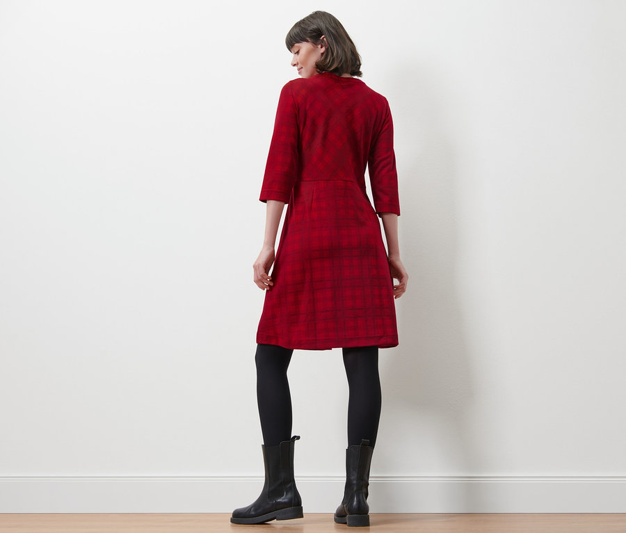 Eine Frau steht mit dem Rücken zur Kamera und trägt ein kariertes Jacquard-Kleid, eine schwarze Leggings und schwarze Stiefel.