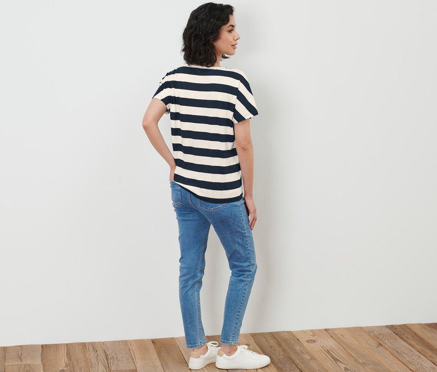 Frau steht seitlich und trägt ein Gestreiftes Blusenshirt, Jeans und Turnschuhe.